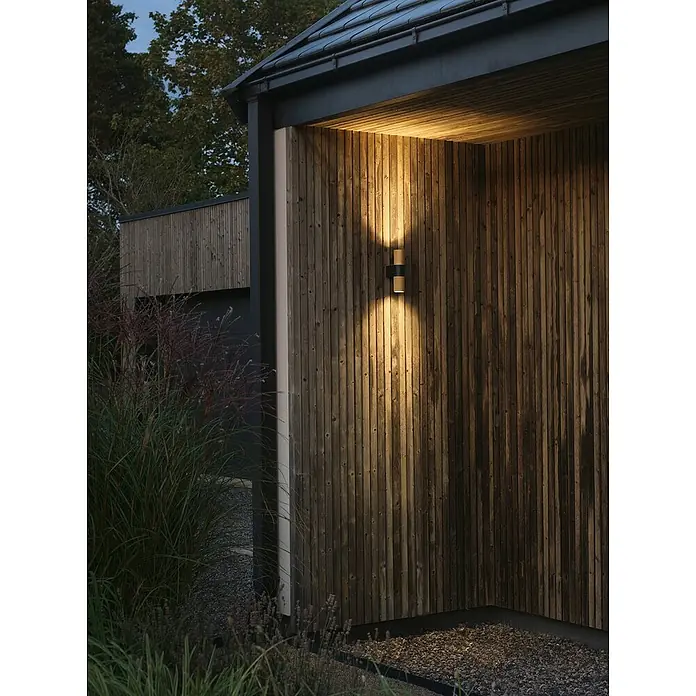 Konstsmide Außenwandleuchte Temi - 14 W, Schwarz/Gold, IP54 - Nahaufnahme einer modernen Wandleuchte an einer Holzwand, die warmes Licht ausstrahlt und eine gemütliche Atmosphäre schafft.