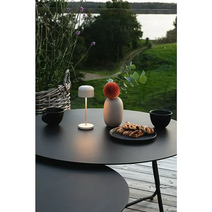Konstsmide Akku-Tischleuchte Lille Mini - 2,5 W, L x B x H: 7,5 x 7,5 x 20 cm, Sand, Mehrfarbig - Ein stimmungsvolles Ambiente mit einer eleganten, kabellosen Tischleuchte auf einem schwarzen Tisch mit Gebäck, perfekt für gemütliche Stunden im Freien.