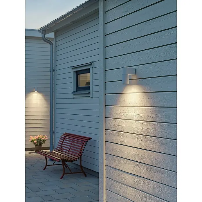 Konstsmide Außenwandleuchte Temi - 7 W, Weiß, IP54 - Bild einer modernen weißen Außenwandleuchte, die ein warmes Licht auf eine Holzfassade wirft. Die Leuchte betont das moderne Design und die einladende Atmosphäre.