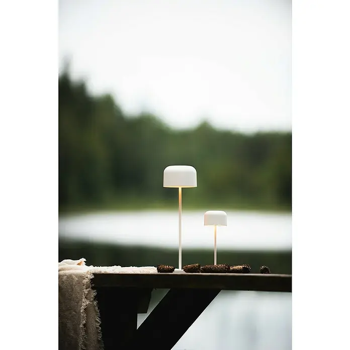 Konstsmide Akku-Tischleuchte Lille - 3,5 W, L x B x H: 11 x 11 x 36 cm, Weiß, Mehrfarbig - Zwei moderne weiße Tischleuchten auf einem Holztisch mit Tannenzapfen. Die Lampen strahlen warmes Licht in die verschwommene Naturkulisse.