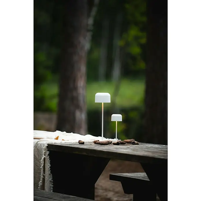 Konstsmide Akku-Tischleuchte Lille - 3,5 W, L x B x H: 11 x 11 x 36 cm, Weiß, Mehrfarbig - Zwei tragbare LED-Tischlampen in Weiß auf einem Holztisch mit Tannenzapfen, ideal für stimmungsvolle Beleuchtung im Freien.