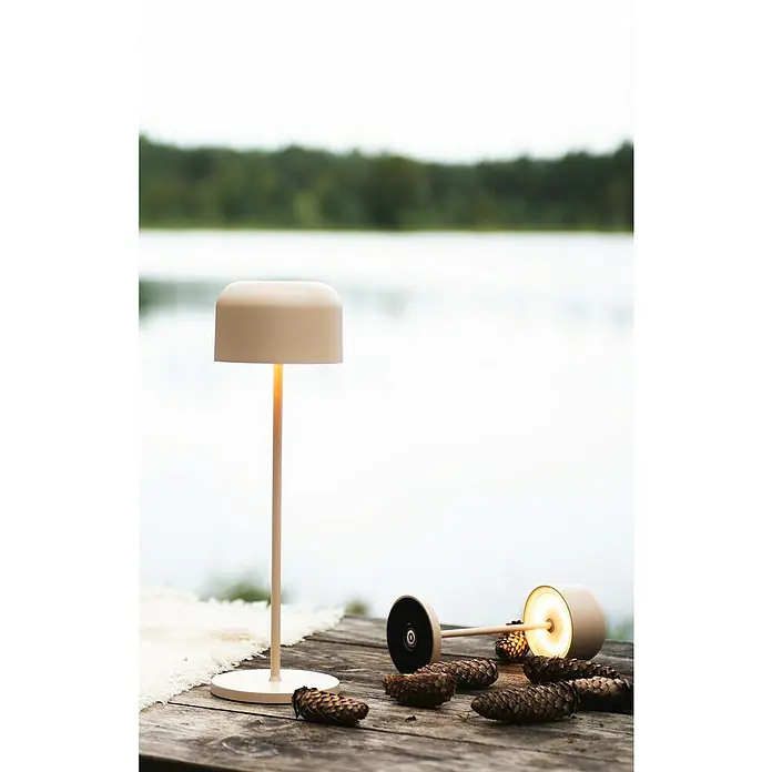 Konstsmide Akku-Tischleuchte Lille - 3,5 W, L x B x H: 11 x 11 x 36 cm, Sand, Mehrfarbig - Stimmungsvolle Szene mit zwei modernen Tischleuchten in Beigetönen und Tannenzapfen auf einer Holzoberfläche vor einem See.
