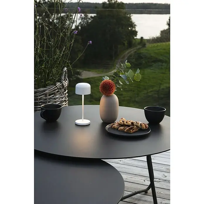 Konstsmide Akku-Tischleuchte Lille Mini - 2,5 W, L x B x H: 7,5 x 7,5 x 20 cm, Weiß, Mehrfarbig - Eine stimmungsvolle Szene mit einer modernen, weißen Tischleuchte, die ein sanftes Licht auf einer schwarzen Tischplatte spendet.