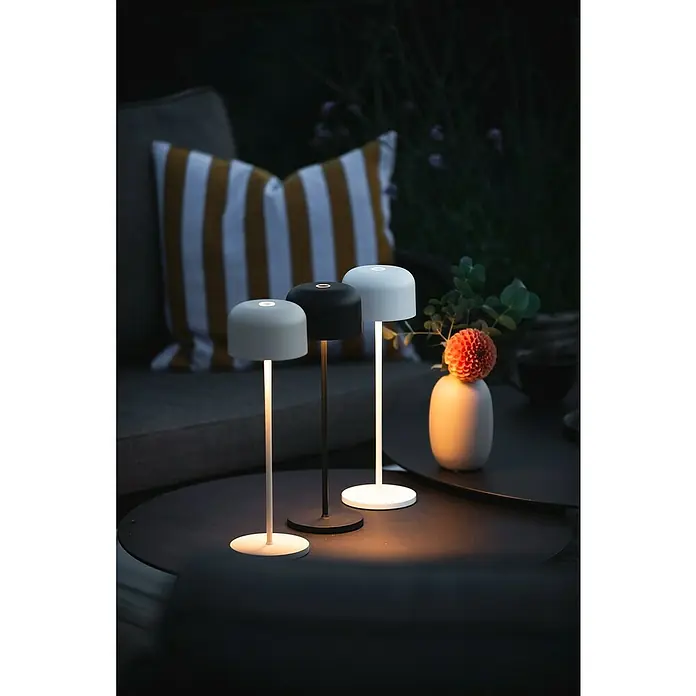 Konstsmide Akku-Tischleuchte Lille - 3,5 W, L x B x H: 11 x 11 x 36 cm, Schwarz, Mehrfarbig - Drei moderne Tischleuchten in Grau, Schwarz und Weiß erhellen einen dunklen Tisch, daneben eine Vase mit Blume.