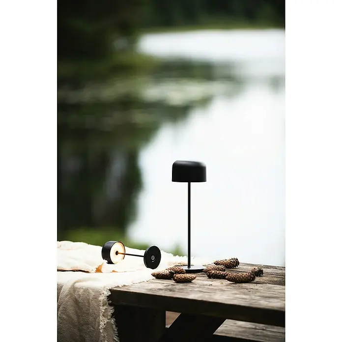 Konstsmide Akku-Tischleuchte Lille - 3,5 W, L x B x H: 11 x 11 x 36 cm, Schwarz, Mehrfarbig - Zwei stilvolle Akku-Tischleuchten in Schwarz und verschiedene Tannenzapfen auf einem Holztisch mit einer weißen Tischdecke.
