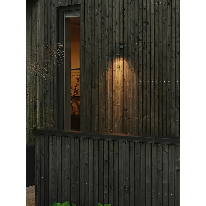 Konstsmide Außenwandleuchte Temi - 7 W, Schwarz, IP54 - Moderne Außenwandleuchte aus schwarzem Metall beleuchtet eine dunkle Holzwand und schafft eine warme Atmosphäre im Außenbereich.