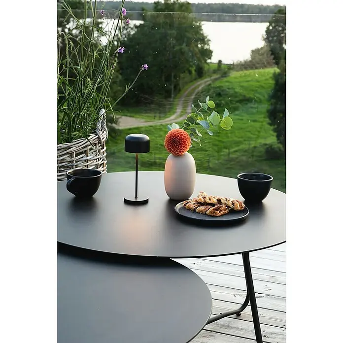 Konstsmide Akku-Tischleuchte Lille Mini - 2,5 W, L x B x H: 7,5 x 7,5 x 20 cm, Schwarz, Mehrfarbig - Ein moderner Tisch mit Lampe, Gebäck und Tassen auf einer Terrasse mit Blick auf die Natur.