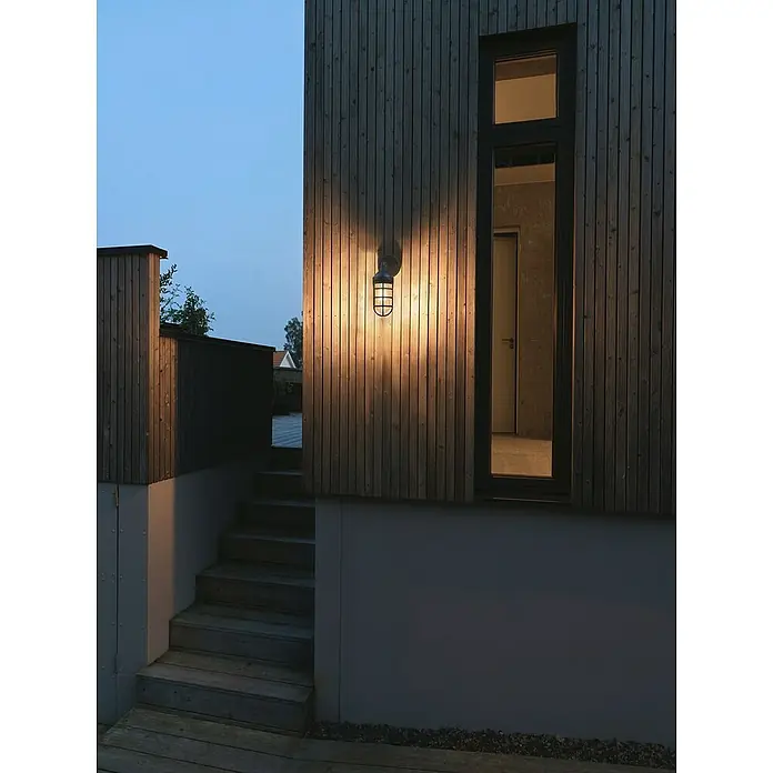 Konstsmide Außenwandleuchte Olbia - 20 W, Schwarz, IP44 - Außenansicht eines Gebäudes mit einer stilvollen Außenlampe, die ein warmes Licht ausstrahlt. Die Fassade besteht aus dunklem Holz und hat ein Fenster.