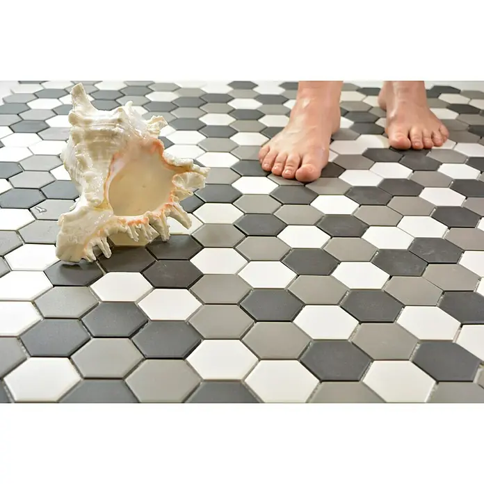 Mosaikfliese Hexagon Mix CU HX150 - 32,5 x 28,1 cm, Weiß/Schwarz/Grau, Matt - Nahaufnahme eines Muschelgehäuses auf einem Boden mit sechseckigen Fliesen in Grau-, Schwarz- und Weißtönen und Füßen im Hintergrund.