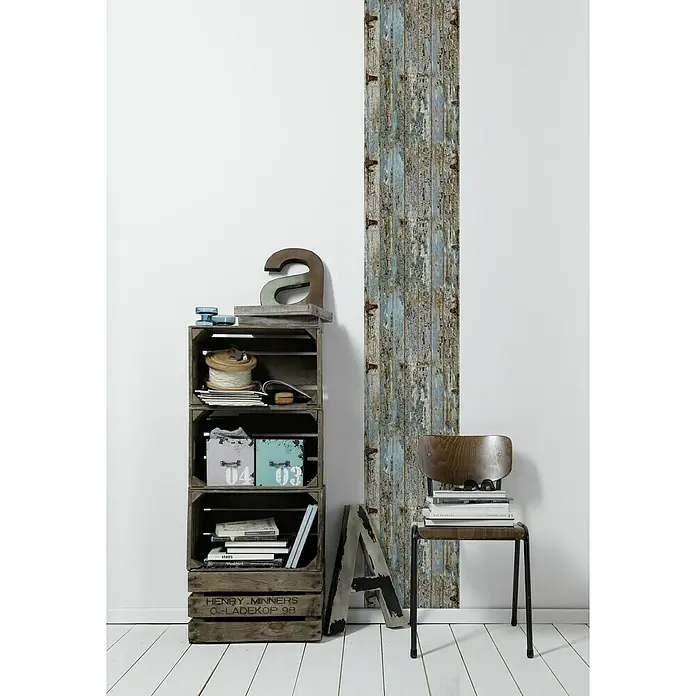 A.S. Création Designpanel pop.up - Holz Vintage, Grau/Blau/Braun, 35 cm x 2,5 m - Dekoratives Regal aus Holzkisten mit Deko-Elementen und ein Stuhl mit Büchern, vor einer Wand mit Holzpaneelen.