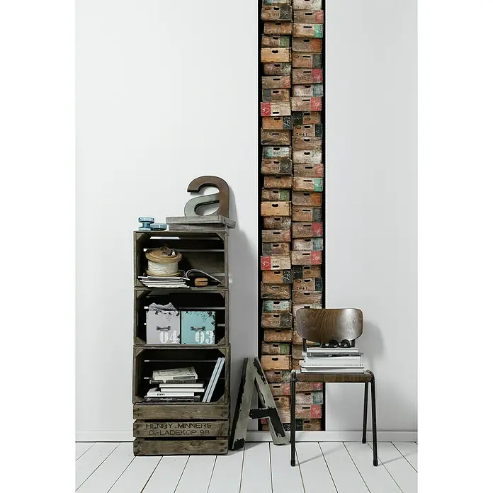 A.S. Création Designpanel pop.up - Kisten, Braun/Schwarz/Bunt, 35 cm x 2,5 m - Regal aus Holzkisten neben einem Stuhl mit Büchern und einem Dekorbuchstaben.