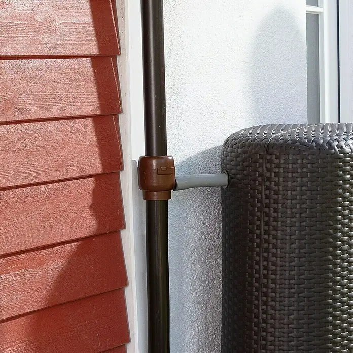 Garantia Füllautomat Mini - Braun, Passend für: Fallrohre von DN 50 - 60 - Nahaufnahme einer Regenwassertonne aus Rattan, verbunden mit einem Fallrohr an einer Hauswand. Das System ist auf Wassergewinnung ausgelegt.