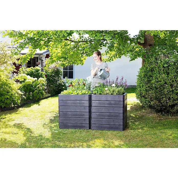 Garantia Hochbeet Ergo Quadro M - 80 x 60 x 25 cm, Kunststoff, Anthrazit, Holzoptik - Eine Frau gießt Pflanzen in einem hölzernen Hochbeet im Garten. Das Beet ist mit Gemüse und Blumen bepflanzt und steht auf einem Rasen.