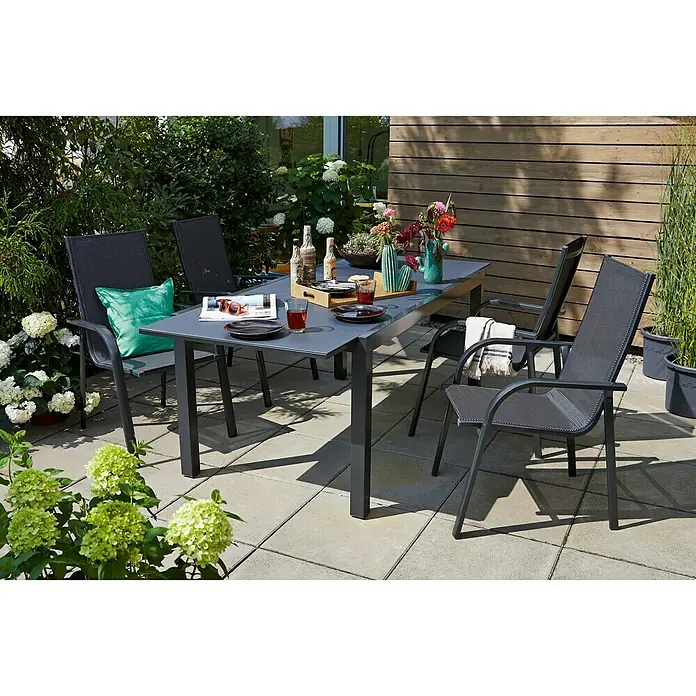 Sunfun Maja Gartenmöbel-Set I - 5 -tlg., Glas/Textilen, Anthrazit - Ein grauer Gartentisch mit vier passenden Stühlen steht auf einer Terrasse, umgeben von üppigem Grün. Perfekt für Mahlzeiten im Freien.