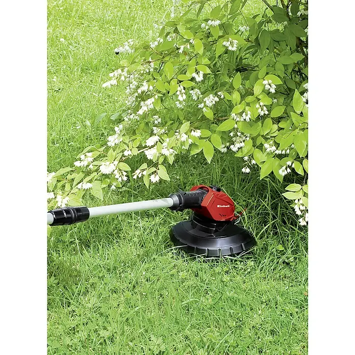 Einhell PXC 18V Recortabordes de batería GE-CT 18 Li-Solo - 18 V, Iones de litio, Sin batería - Un cortacésped inalámbrico Einhell rojo está sentado en la hierba debajo de un arbusto en flor. El dispositivo está listo para usar en el jardín.