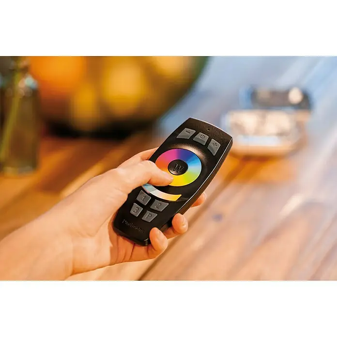 Paulmann Smart Home ZigBee Mando a distancia - Alcance radiodifusión: 50 m, 2,4 GHz - Una mano sujeta un mando a distancia Paulmann para el control de la iluminación con rueda de colores y botones sobre una mesa de madera.