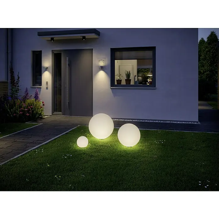 Paulmann Plug & Shine Aplique para exterior LED Globe - 2,8 W, IP67, Blanco cálido - Esferas de jardín iluminadas de varios tamaños adornan el césped frente a una casa moderna, creando un ambiente atmosférico.