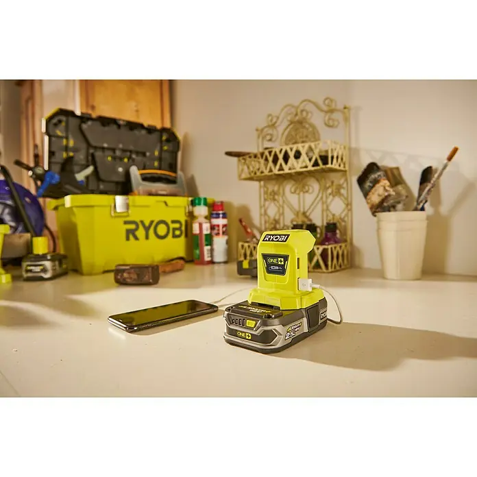 Ryobi ONE+ Adaptador USB R18USB - 18 V, Sin batería - Primer plano de un cargador Ryobi ONE+ con un smartphone conectado por USB en una mesa.