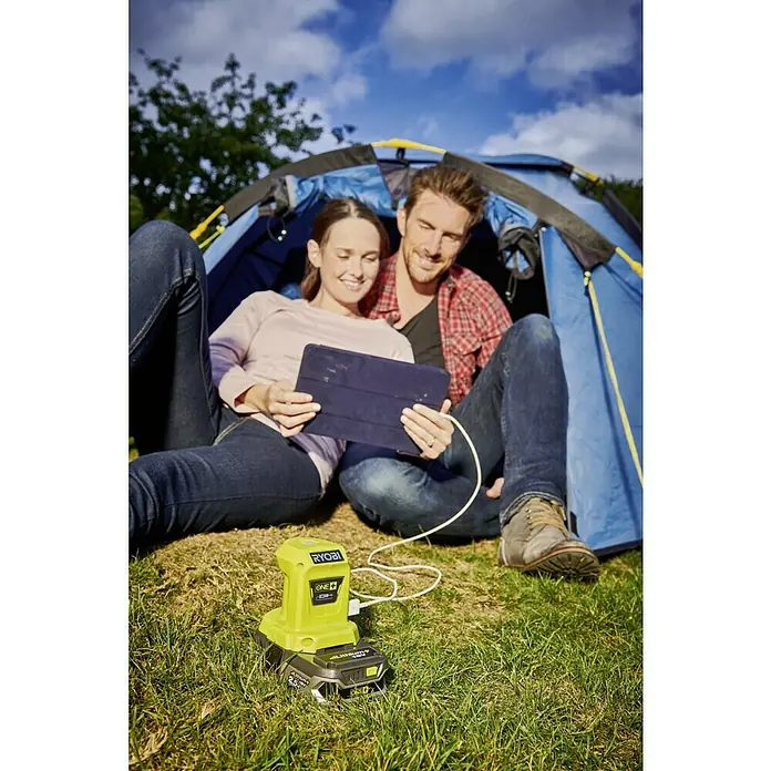 Ryobi ONE+ USB-Adapter - 18 V, Ohne Akku - Ein Paar zeltet und nutzt ein RYOBI ONE+ Ladegerät, um ihr Tablet aufzuladen. Das grüne Ladegerät steht im Vordergrund auf dem Rasen.