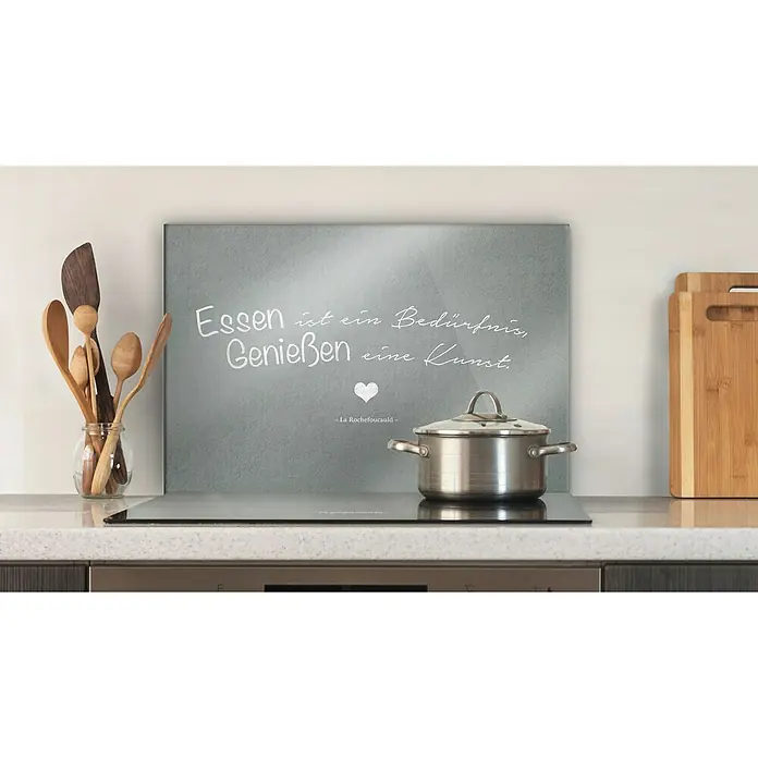 CUCINE Küchenrückwand - Essensgenuss, 60 x 40 cm, Stärke: 6 mm, Einscheibensicherheitsglas (ESG) - Das Bild zeigt ein Kochfeld mit einem Topf darauf, vor einer Tafel mit der Aufschrift "Essen ist ein Bedürfnis, Genießen eine Kunst".