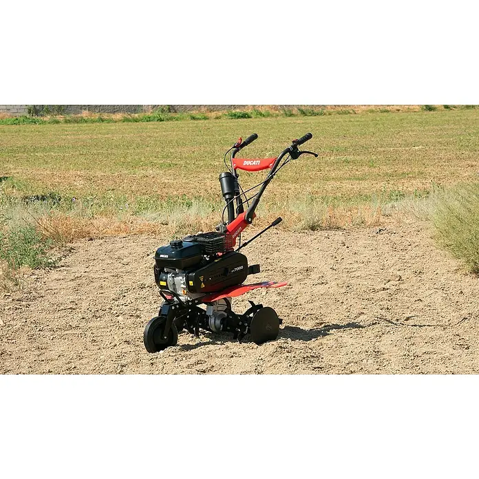 Ducati Azada de gasolina DTL7000 - 4,3 kW, 4 tiempos, Recomendación máxima de superficie: 1.500 m² - Una motoazada roja Ducati está de pie en un campo. La motoazada está diseñada para el cultivo del suelo en jardines y mejora la salud del suelo.