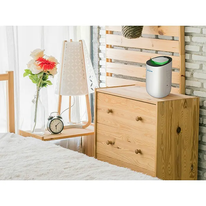 Purline Deshumidificador eléctrico Dryos 1 - 23 W, L x An x Al: 14,6 x 14,6 x 22,5 cm, Blanco - Purificador de aire sobre una cómoda de madera junto a una lámpara de noche y flores. Ideal para aire limpio en tu dormitorio.