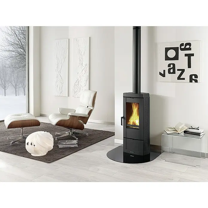 La Nordica Kaminofen Candy - 8,7 kW, Raumheizvermögen: 206 m³, Material Verkleidung: Stahl, Schwarz - Bild eines modernen Kaminofens mit Glasablage, Sessel und winterlicher Aussicht. Der Kaminofen strahlt Wärme und Eleganz aus.