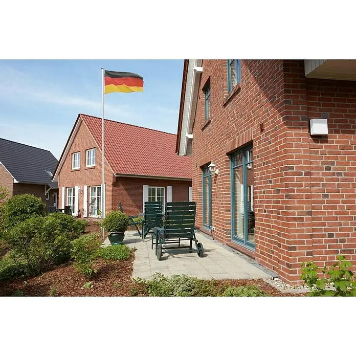 Fahnenmast - Höhe: 6,2 m, Aluminium - Deutsche Flagge weht vor Backsteinhäusern. Die rot-gold-schwarze Flagge schmückt einen sonnigen Außenbereich mit Gartenmöbeln und Steinplatten.