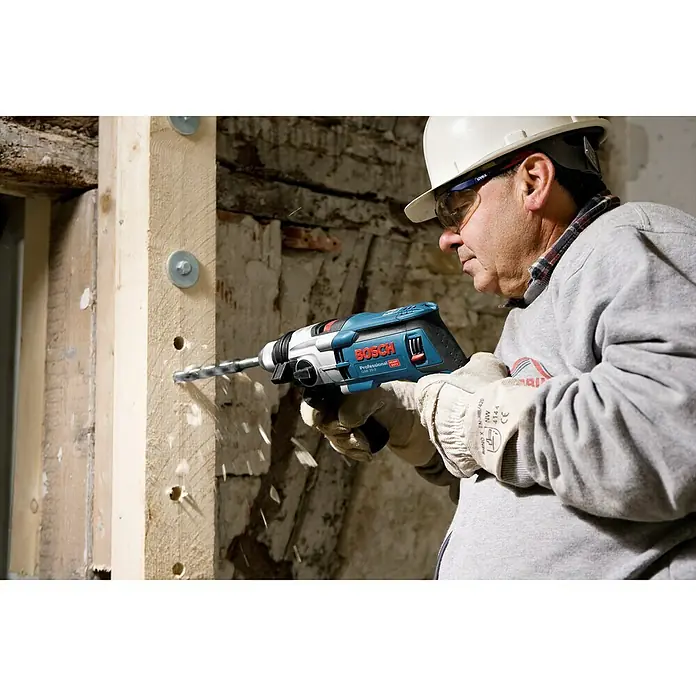 Bosch Professional Schlagbohrmaschine GSB 20-2 - 850 W, Schnellspannbohrfutter, Ohne Steinbohrer-Set - Ein Bauarbeiter mit Schutzhelm und Brille bohrt mit einer blauen Bosch Professional Bohrmaschine ein Loch in einen Holzbalken.