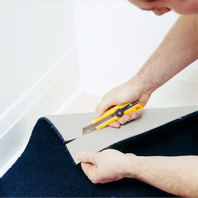 Olfa Cuttermesser L-1 - Gelb/Schwarz - Eine Nahaufnahme von jemandem, der mit einem gelben Cuttermesser ein Stück marineblauen Teppich auf einem hellgrauen Untergrund zuschneidet.