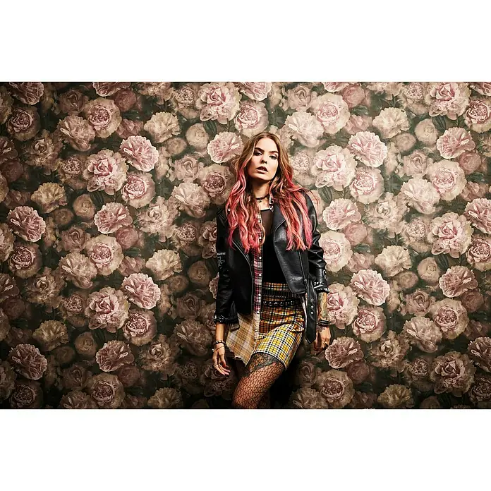 A.S. Création Neue Bude 2.0 ED II Vliestapete Barockrose - Altrosa, Floral, 10,05 x 0,53 m - Eine stilvolle Frau mit rosa Haarsträhnen, gekleidet in eine Lederjacke und einen karierten Rock, posiert vor einem floralen Hintergrund.