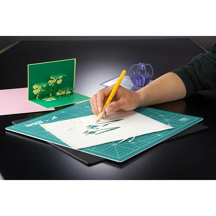 Olfa Präzisionsmesser AK-1Skalpellklinge, 6 -tlg. - Jemand schneidet mit einem Bastelmesser ein filigranes Blumenmuster auf einer grünen Schneidematte, um kreative Papierkunst zu schaffen.