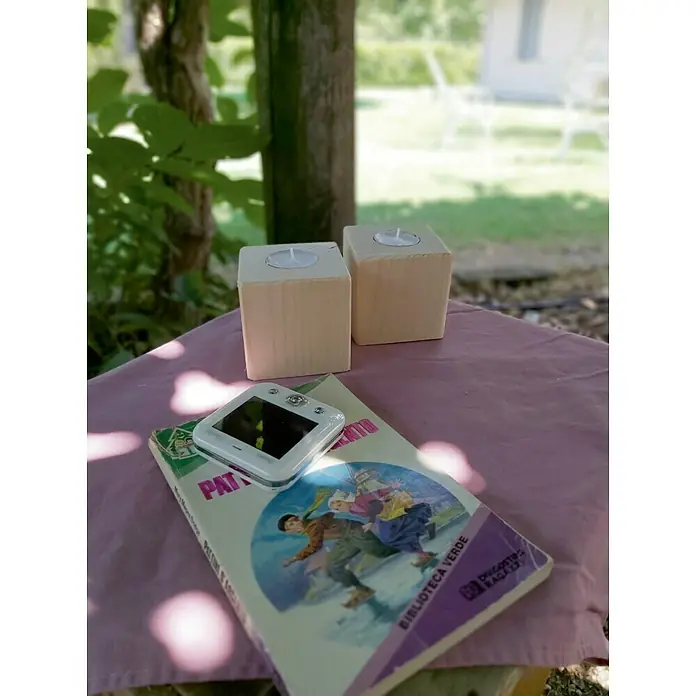 Portavelas - 8 x 8 x 8 cm, Madera, Natural - Primer plano de dos portavelas de cubo de madera, un lector electrónico y un libro sobre una manta morada al aire libre.