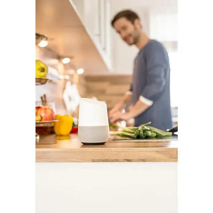 Schellenberg Funk-Rohrmotor Premium 6 MiniMax. Rollladenfläche: 4 m² bei Kunststoffrollläden, Einbaulänge: 570 mm, Zugkraft: 15 kg - Bild eines Google Home Smart Speakers auf einer Küchentheke, mit einem Mann im Hintergrund, der Gemüse zubereitet.