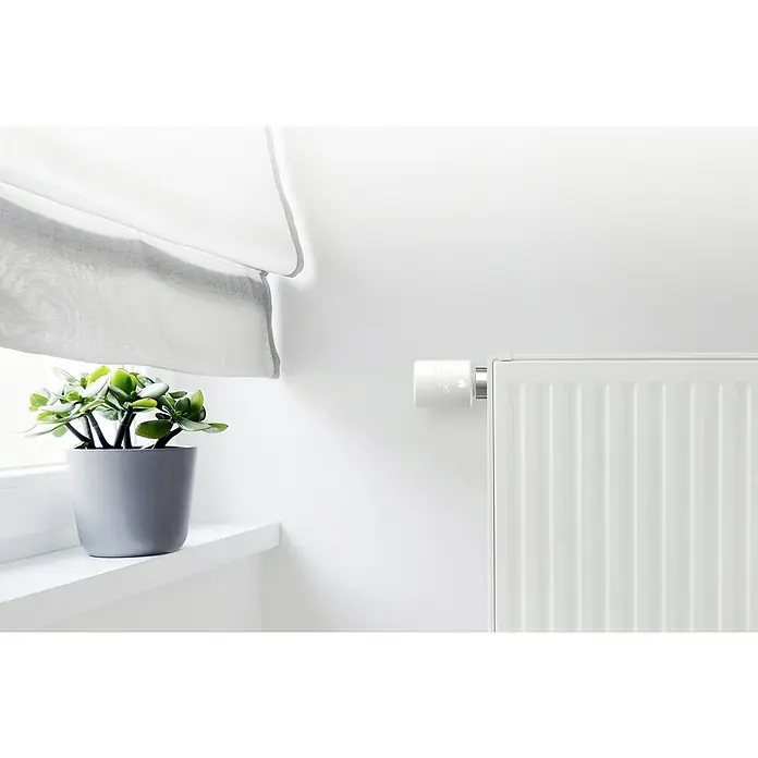 Tado Smartes Termostato de radiador Starter Kit V3+IN - 2 pzs. - Primer plano de un termostato de radiador inteligente conectado a un radiador junto a un alféizar con una planta.