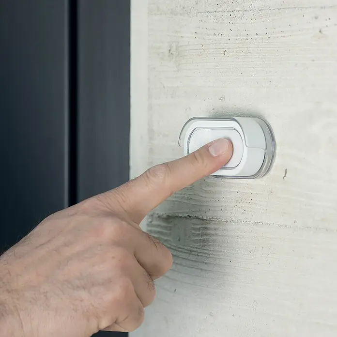 Philips Funk-Gong-Set WelcomeBell 300 Color - Reichweite Funk: 300 m, IP65, Kunststoff - Eine Hand drückt einen weißen Klingelknopf mit durchsichtigem Schutz an einer Betonwand, neben einer dunklen Tür.