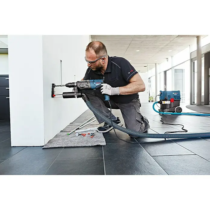 Bosch Professional Absaugvorrichtung GDE 16 Plus - Passend für: Bosch Professional Bohrhämmer 2 & 3 kg - Ein Handwerker bohrt mit einer blauen Bosch-Bohrmaschine ein Loch in eine weiße Wand, während ein Staubsauger den Staub absaugt.