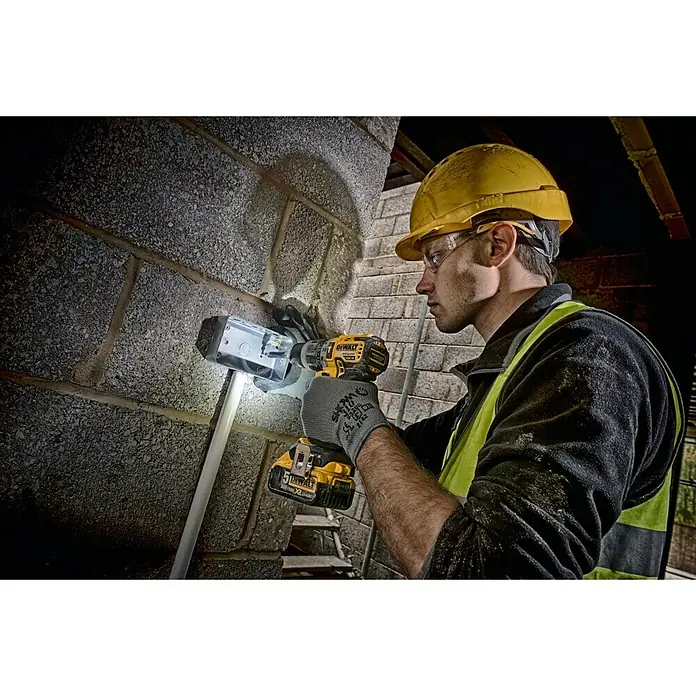 DeWalt XR 18V Taladro atornillador percutor de batería - 18 V, Número máx. de impactos: 34.000 /min, Sin batería, Niveles de par de giro: 15 ud. - Un trabajador de la construcción asegura una caja de conexiones gris a una pared usando un taladro inalámbrico amarillo DeWalt. Lleva gafas de seguridad y casco amarillo.