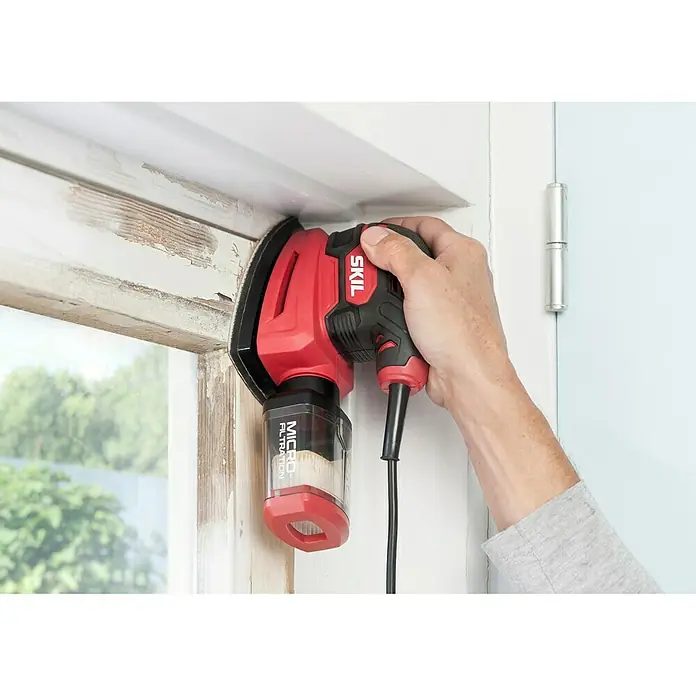 Skil Elektro-Multischleifer 7260 AA - 100 W, Leerlaufschwingzahl: 24.000 U/min, Durchmesser Schwingkreis: 1,6 mm - Nahaufnahme eines roten SKIL-Detail-Schleifers in Aktion, der an einem Fensterrahmen verwendet wird, mit Blick auf den integrierten Staubbehälter.