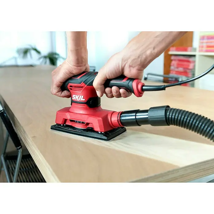 Skil Elektro-Schwingschleifer 7362 AA - 280 W - Nahaufnahme eines roten SKIL-Schleifgeräts, das von zwei Händen auf einer Holzoberfläche gehalten wird, mit angeschlossenem Staubsaugerschlauch.