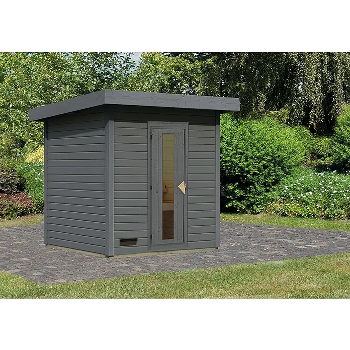 Karibu Saunahaus Hytti - Ohne Ofen, Terragrau, L x B x H: 196 x 196 x 228 cm - Eine graue Gartensauna mit Holztür und kleinem Fenster steht auf einem gepflasterten Bereich. Im Hintergrund üppiges Grün.