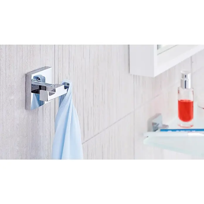 Tesa Deluxxe Handtuchhaken - 2-armig, Chrom, Glänzend - Nahaufnahme eines verchromten Handtuchhalters mit blauem Handtuch im Badezimmer, darüber ein weißer Schrank und eine Flasche mit roter Flüssigkeit.