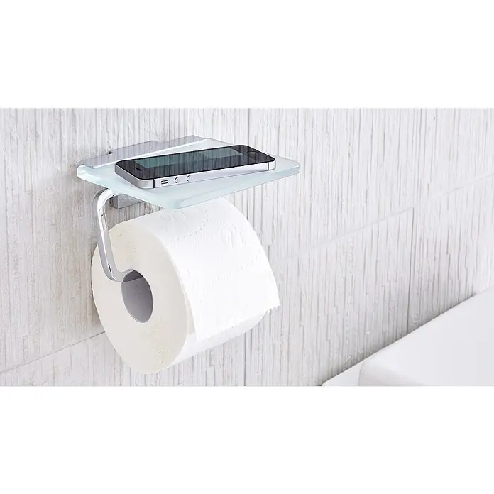 Tesa Deluxxe Papierrollenhalter - Mit Ablage, Chrom, Glänzend - Toilettenpapierhalter mit Ablage aus Glas, auf der ein Smartphone liegt, an einer weißen Wand montiert.