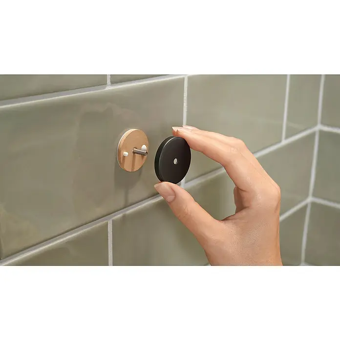 Tesa Aluxx Cesta de baño angular - L x An x Al: 16,3 x 16,3 x 26,2 cm, Negro, Recubrimiento en polvo - Una mano sostiene una pieza redonda y negra lista para fijarse a un soporte de madera con un tornillo, ya fijado a una pared alicatada.