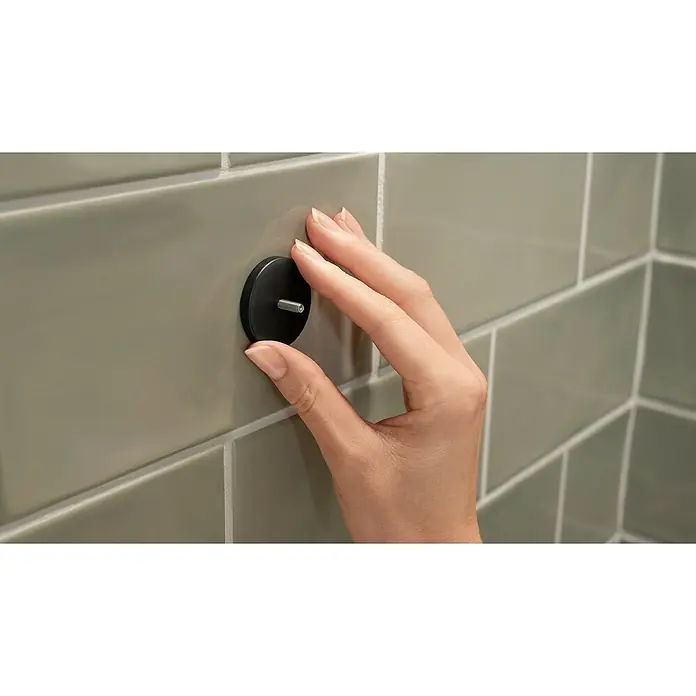 Tesa Aluxx Cesta de baño angular - L x An x Al: 16,3 x 16,3 x 26,2 cm, Negro, Recubrimiento en polvo - Una mano está asegurando un soporte de pared redondo negro a una pared de azulejos de color gris claro con un tornillo en el centro.