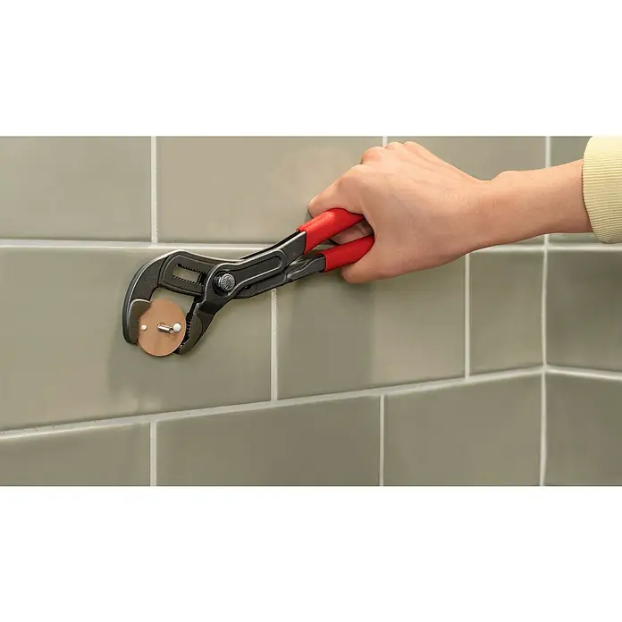 Tesa Aluxx Cesta de baño angular - L x An x Al: 16,3 x 16,3 x 26,2 cm, Negro, Recubrimiento en polvo - Una llave de tubo con mangos rojos sujeta un accesorio de baño marrón contra un fondo de azulejos grises, ideal para reparaciones.