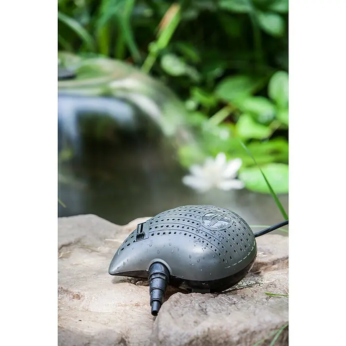 Heissner Filterpumpe - Fördermenge pro Std.: 3.300 l - Nahaufnahme einer Heissner-Gartenpumpe auf einem Stein mit Teich und Pflanzen im Hintergrund.