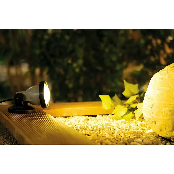 Heissner Teichbeleuchtung - 4,5 W, Warmweiß - Dämmerungsaufnahme einer hellen Gartenleuchte auf einer Holzterrasse, die warmes Licht auf Kieselsteine und eine Kugel wirft.