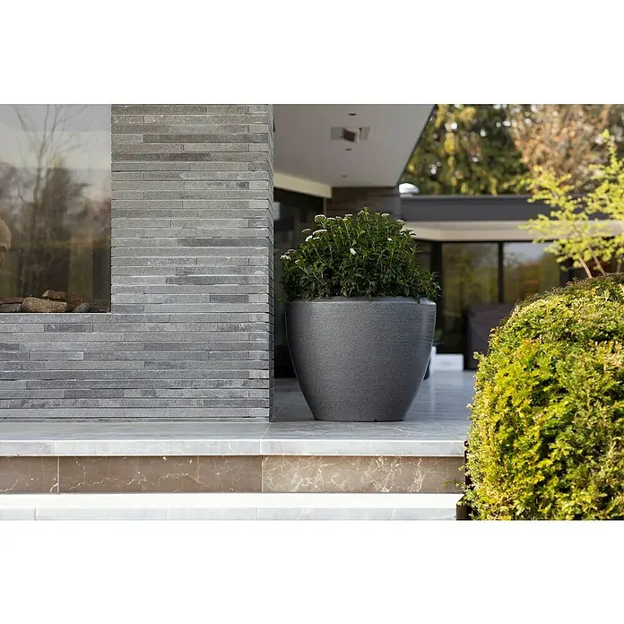 Capi Europe Übertopf rund Arc Granite - Außenmaß (Ø x H): 34 x 25 cm, Anthrazit, Kunststoff, Matt - Ein großer, grauer Pflanzkübel mit üppigem Grün steht vor einer modernen Fassade, die mit grauen Steinplatten verkleidet ist.
