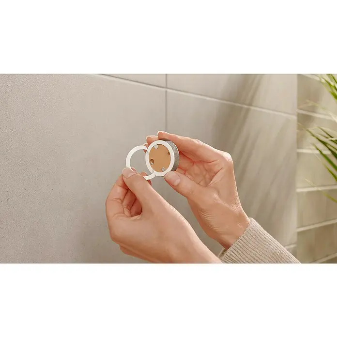 Tesa Moon Handtuchhaken - 2-armig, Graphit, Matt - Die Hände einer Person halten ein rundes, silbernes Gerät mit einer abnehmbaren, weißen Scheibe vor einer hellen Wand.